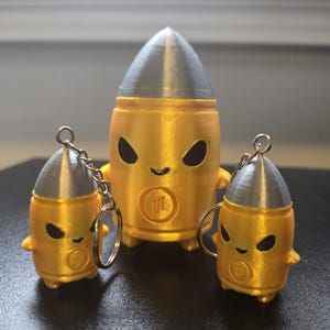 Könnte beinhalten: Drei patronenförmige Figuren mit goldenen Körpern und silbernen Spitzen. Jede Figur hat schwarze mandelförmige Augen, ein kleines Lächeln und ein kreisförmiges Emblem auf der Vorderseite. Zwei der Figuren sind kleiner und haben Metallschlüsselanhänger.