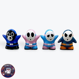 Op de afbeelding: Vier kleine karakterfiguurtjes staan op een rij. De eerste is blauw en zwart met een rond masker. De tweede is roze en blauw met een ovaal masker. De derde is lichtblauw en roze met een lachend masker. De vierde is donkerblauw en oranje.