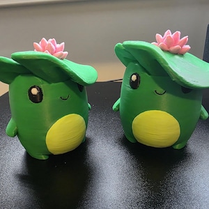 Könnte beinhalten: Zwei grüne, froschförmige Figuren mit gelben Bäuchen, schwarzen Augen und einem kleinen Lächeln. Jeder Frosch trägt ein großes grünes Seerosenblatt auf dem Kopf, das von einer kleinen rosa Lotusblüte gekrönt wird. Die Figuren haben eine matte Oberfläche.