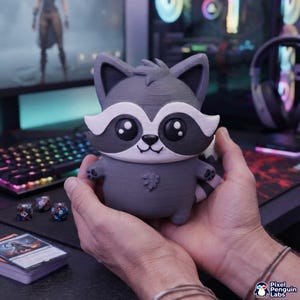 Könnte beinhalten: Eine 3D-gedruckte Waschbär-Figur mit grauem Körper, weißen Gesichtsmarkierungen, großen schwarzen Augen und einer kleinen schwarzen Nase. Die Figur hat einen gestreiften Schwanz, kleine Pfoten und eine texturierte Oberfläche als kompaktes Sammlerstück.