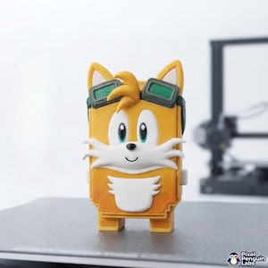 Könnte beinhalten: Eine 3D-gedruckte rechteckige Figur des Charakters Tails aus Sonic the Hedgehog mit gelbem Fell, weißer Brust und weißen Wangenbüscheln, einer kleinen schwarzen Nase und einer dunkelgrünen Schutzbrille auf der Stirn.