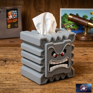 Op de afbeelding: Een grijze, kubusvormige tissueboxhouder ontworpen als een Thwomp-personage. De box heeft een getextureerd, blokkerig oppervlak met een opvallend, boos gezicht met rode ogen en witte tanden. Een wit tissue steekt uit de opening aan de bovenkant.
