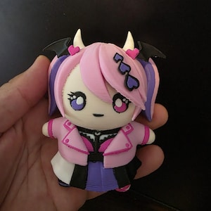 Könnte beinhalten: Eine kleine, dreidimensionale Figur eines Charakters mit rosa Haaren, kleinen weißen Hörnern und schwarzen fledermausartigen Flügeln. Die Figur trägt eine rosa Jacke über einem schwarz-violetten Outfit und hat herzförmige Haaraccessoires.