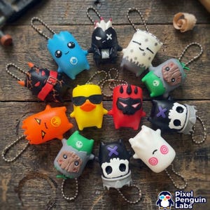 Peut inclure: Une collection de douze petits porte-clés personnages colorés avec des chaînes à billes en métal. Les figurines présentent divers designs, dont un canard jaune avec des lunettes de soleil, un crâne noir, une créature bleue et un fantôme blanc.