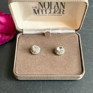 May include: A pair of silver-toned stud earrings featuring a central oval-cut clear stone surrounded by a halo of smaller baguette-cut stones. The earrings are presented in a beige velvet-lined box labeled The Nolan Miller Glamour Collection.