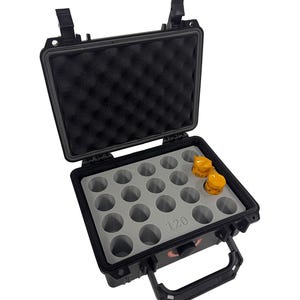 Puede incluir: Maletín rígido negro con interior de espuma gris y 24 compartimentos. El maletín tiene un asa y dos cierres. El maletín está abierto y hay tres contenedores amarillos en el interior.