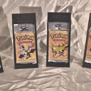 May include: Four sealed Pokémon trading card booster packs, each displayed in a vertical black plastic stand. The packs feature illustrations of Charizard, Aerodactyl, Zapdos, and Lapras, with the Pokémon logo printed at the top of each pack.