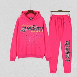 Op de afbeelding: Een felroze hoodie en bijpassende joggingbroek. De hoodie heeft een zwarte afbeelding met de tekst "spider" en "never the same", omringd door een spinnenweb en kleurrijke sterren. De broek heeft dezelfde tekst verticaal op de zijkant gedrukt.