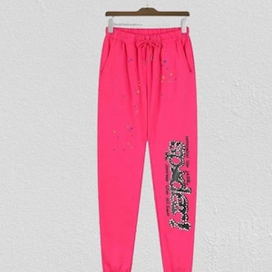 Op de afbeelding: Felroze joggingbroek met een elastische tailleband met trekkoord, zijzakken en verspreide veelkleurige verfspatten. De linkerpijp is voorzien van een grote, zwarte, met studs versierde afbeelding met de tekst Spider en bijbehorende tekst.