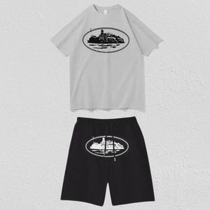 Puede incluir: Un conjunto de camiseta de manga corta gris claro y pantal&oacute;n corto negro. La camiseta presenta un gr&aacute;fico ovalado negro de un barco en el pecho. El pantal&oacute;n corto negro tiene un gr&aacute;fico ovalado blanco del mismo barco con cord&oacute;n en la cintura.