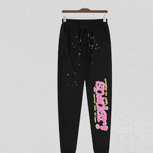 Op de afbeelding: Zwarte joggingbroek met een trekkoord in de taille, veelkleurige verfspatten en een grote roze afbeelding op de linkerpijp met de tekst "spider!" en kleinere gele tekst "nevermind the punk, here's the alien".
