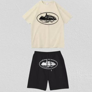 Puede incluir: Un conjunto de camiseta de manga corta color crema y pantalones cortos negros. La camiseta presenta un gr&aacute;fico ovalado negro de un barco en el pecho. Los pantalones cortos negros tienen un gr&aacute;fico ovalado blanco a juego en la pierna y cintura con cord&oacute;n.