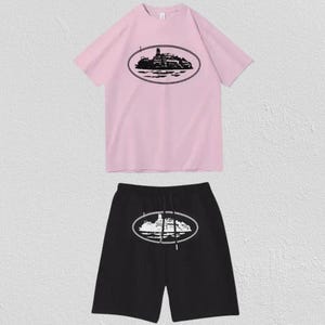 Puede incluir: Un conjunto de camiseta rosa claro de manga corta y pantal&oacute;n corto negro. La camiseta presenta un gr&aacute;fico ovalado negro de un barco en el pecho. El pantal&oacute;n corto negro muestra un gr&aacute;fico ovalado blanco del mismo barco y tiene un cord&oacute;n ajustable.
