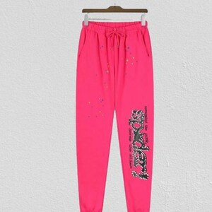 Op de afbeelding: Felroze joggingbroek met een elastische tailleband met trekkoord, zijzakken en verspreide veelkleurige verfspatten. De linkerpijp is voorzien van een grote, zwarte, met studs versierde afbeelding met de tekst Spider en bijbehorende tekst.
