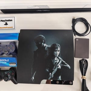 May include: A black PlayStation 3 game console featuring The Last of Us artwork, a boxed purple DualShock 3 controller, a loose black controller, a Seagate external hard drive, a power cable, a USB cable, an AV cable, and a small PlayStation logo sign.