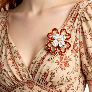 Könnte beinhalten: Eine goldfarbene Brosche in Form eines vierblättrigen Kleeblatts mit roter Emaille an den Blatträndern und einem zentralen Cluster aus klaren, funkelnden Kristallen. Die Brosche ist an einem beigen Kleid mit rotem Blumenmuster befestigt.
