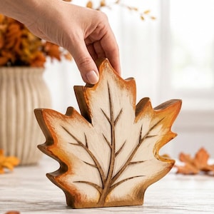 May include: A wooden maple leaf cutout featuring a cream-coloured centre with dark brown carved veins and edges that transition from light orange to deep brown. The leaf stands upright on a flat base and has a distressed, rustic finish.