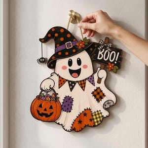 May include: A decorative wooden wall hanging featuring a white ghost wearing a black witch hat with orange polka dots. The ghost holds an orange jack-o'-lantern bucket and a sign that reads BOO!. It is decorated with colourful patches and bunting.