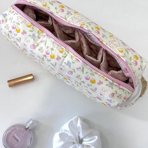 Può includere: Un astuccio rettangolare in tessuto con sfondo bianco e un motivo ripetuto di piccoli tulipani rosa e gialli, foglie verdi e conigli bianchi. L'astuccio presenta una chiusura a zip rosa chiaro, un manico in tessuto abbinato e una fodera rosa.