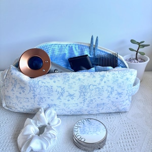 May include: A rectangular fabric pouch with a blue and white toile pattern on the exterior and a blue and white gingham lining. The pouch contains a copper-coloured hair dryer, a black comb, and two blue hair clips. A white scrunchie and a round compact with the text Sulwhasoo are placed in front.