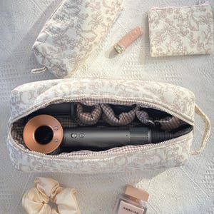 Könnte beinhalten: Eine große rechteckige Kulturtasche mit beigem und weißem Blumenmuster, die einen dunkelgrauen Haartrockner mit kupferfarbener Düse enthält. Ebenfalls enthalten sind ein kleineres passendes Täschchen, ein rechteckiges Reißverschlussetui, ein Haargummi und ein Parfümflakon.