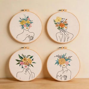 Puede incluir: Cuatro bastidores de bordado circulares de madera con tela blanca y dibujos de líneas negras de figuras humanas. Cada cabeza está reemplazada por un colorido arreglo floral con flores amarillas, rosas y naranjas y hojas de color verde.