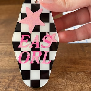 May include: A motel-style keychain with a black and white checkered pattern. The keychain features two pink glittery stars and the text "EAS ORI" in a pixelated pink font. It is attached to a silver metal split ring for keys.