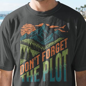 Könnte beinhalten: Ein anthrazitfarbenes T-Shirt mit einem Grafikdruck von einem Gebirge, einem Wald und einer Sonne. Der Text "DON'T FORGET THE PLOT" ist in großen, fetten, orangefarbenen und grünen Buchstaben über den unteren Teil des Berglandschaftsmotivs gedruckt.