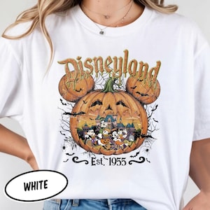 May include: A white t-shirt featuring a graphic of three orange pumpkins arranged in a Mickey Mouse shape. The central pumpkin contains a scene with cartoon characters and a castle. The text reads Disneyland and Est. 1955, with spiderwebs and bats.