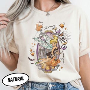 Könnte beinhalten: Ein cremefarbenes T-Shirt mit einer Grafik von Tinker Bell in einem verzierten violetten Rahmen. Das Design zeigt einen Kürbis, Zuckermais, Fledermäuse, ein Spinnennetz sowie den Text "SPOOKY" und "TINKER BELL" mit einem Mickey-Ballon.