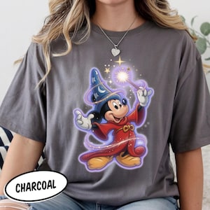 May include: A charcoal gray short-sleeve t-shirt featuring a graphic of Mickey Mouse dressed as a wizard in a red robe and blue star-patterned hat, holding a glowing wand with sparkling light effects and purple magical swirls surrounding the character.