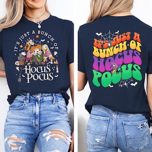 May include: A navy blue t-shirt featuring a front graphic of cartoon characters and the text "IT'S JUST A BUNCH OF HOCUS POCUS" and a back graphic with the same text in large, colorful, wavy letters over a spiderweb and witch hat design.