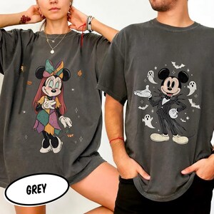 May include: Two charcoal grey t-shirts featuring Disney characters. The left shirt displays a Sally-inspired character with a patchwork dress and bow. The right shirt features a Jack Skellington-inspired character in a pinstripe suit with ghosts.