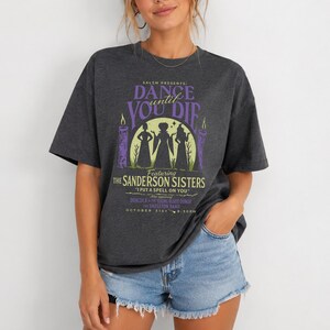 May include: A dark charcoal gray short-sleeve t-shirt featuring a graphic print with the text SALSM PRESENTS DANCE UNTIL YOU DIE, FEATURING THE SANDERSON SISTERS, I PUT A SPELL ON YOU, DRACULA, THE YOUNG BLOOD DONOR, THE SKELETON BAND, OCTOBER 31ST.