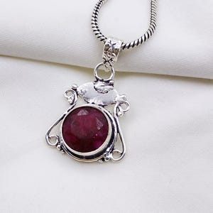 Könnte beinhalten: Eine silberne Anhängerkette mit einem runden, facettierten, tiefroten Edelstein, der in einen dekorativen Metallrahmen mit komplizierten Schnörkeldetails gefasst ist. Der Anhänger hängt an einer silbernen Schlangenkette mit poliertem Finish.