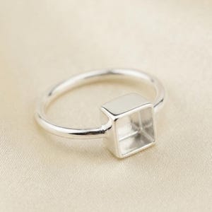 Puede incluir: Un anillo de tono plateado con una banda lisa y pulida, que presenta un engaste cuadrado hueco y prominente en la parte superior. La superficie metálica tiene un acabado brillante y reflectante, y el componente cuadrado está abierto.