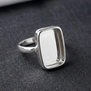 Puede incluir: Un anillo de tono plateado que presenta un engaste rectangular grande con esquinas redondeadas. El anillo tiene un acabado metálico liso y pulido, con una banda ancha y sólida que se estrecha ligeramente hacia atrás, diseñado para una piedra.