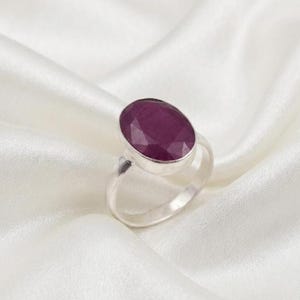 Könnte beinhalten: Ein Silberring mit einem großen, ovalen, tief magentafarbenen Edelstein, der in einer glatten Metallfassung sitzt. Der Ring besteht aus poliertem Silber und der Stein hat eine facettierte Oberfläche, die das Licht reflektiert und glänzt.