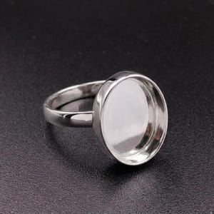 May include: A silver-toned metal ring featuring a smooth, circular bezel setting designed for a cabochon or inlay. The ring has a polished, reflective finish and a simple, solid band that connects to the round, cup-like setting at the top.