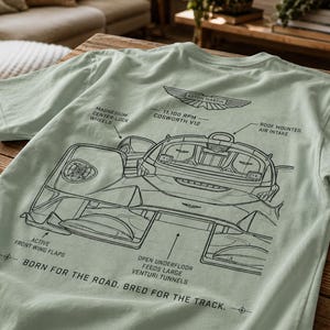 以下が含まれることがあります： ライトセージグリーンのTシャツで、アストンマーティンの車の黒い線画が描かれています。グラフィックには、Magnesium Center-Lock Wheels、11,100 RPM Cosworth V12といった技術的なラベルと、Born for the road. Bred for the track.というテキストが含まれています。