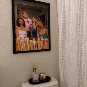 May include: A rectangular black-framed wall print featuring a photograph of four individuals standing behind a row of colourful books. The image shows people in casual clothing, including a pink graphic t-shirt and a blue tank top, in an indoor setting.