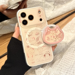 May include: A clear smartphone case with a beige backing featuring various line-art cat illustrations, the text "Kitty" and "cat's Daily", and a pink cat-shaped phone grip attached to the right side. The case has a camera cutout with gold-toned rings.