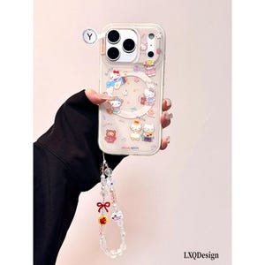 May include: A clear phone case featuring Hello Kitty character graphics, a circular magnetic ring, and a decorative beaded wrist strap with a red bow, a yellow apple charm, and a white heart charm. The case includes the text HELLO KITTY.