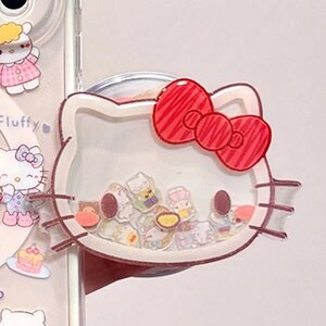 May include: A clear, cat-shaped phone grip featuring a red bow and filled with small, colorful, loose character-themed charms inside. The grip is attached to a transparent phone case decorated with additional small character stickers.