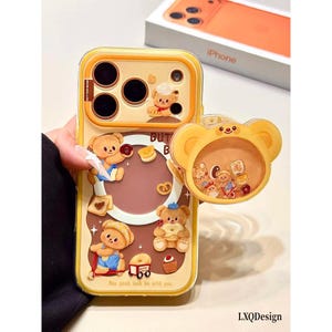 Pode incluir: Uma capa de telemóvel amarela e castanha com ursos de desenhos animados, pastelaria e o texto Butter Bear. Inclui um suporte circular em forma de urso com uma janela transparente contendo pequenos berloques flutuantes em forma de urso e comida.