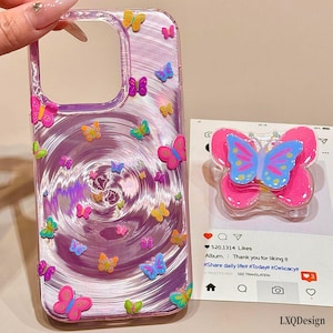 Può includere: Una cover per telefono trasparente con un motivo a vortice viola e vari piccoli adesivi a farfalla colorati, abbinata a un supporto per telefono a forma di farfalla nei colori rosa, blu e giallo su una scheda di interfaccia social media.