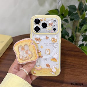 Pode incluir: Uma capa de telemóvel transparente com rebordo amarelo, com ilustrações de coelhos e ursos, gráficos de pão e o texto "Life is like bread just made from the oven." Um suporte de telemóvel em forma de torrada está fixado na parte de trás.