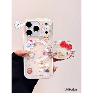 May include: A clear phone case featuring Hello Kitty graphics, including a character in an airplane, a bear, and various small icons. The case includes a circular magnetic ring and a detachable Hello Kitty head-shaped phone grip with a red bow.