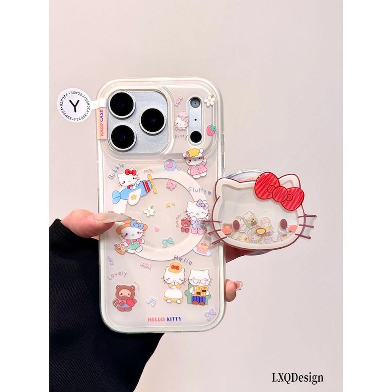 May include: A clear phone case featuring Hello Kitty graphics, including a character in an airplane, a bear, and various small icons. The case includes a circular magnetic ring and a detachable Hello Kitty head-shaped phone grip with a red bow.