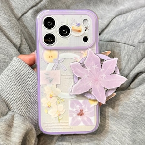 May include: A light purple smartphone case featuring a collage of floral and food illustrations, including a slice of cake, an apple, and white orchids. A large, light purple, star-shaped flower grip is attached to the back of the case.